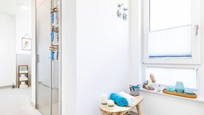 A modern bathroom with a glass shower door, a small wooden stool with toiletries, and a window with a view of the outside.