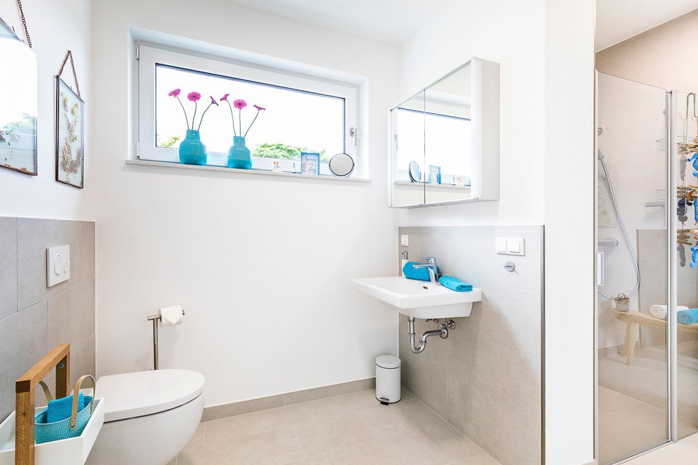 A modern bathroom with a toilet, sink, and shower. There is a window with flowers, a mirror, and a towel rack.