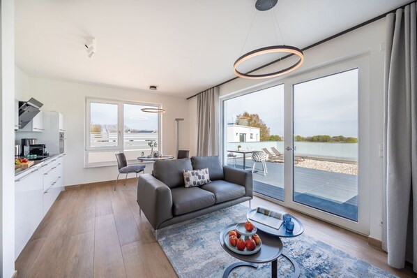Una sala de estar moderna con un sofá gris, una mesa de comedor y un tapete azul, con vista a una masa de agua a través de puertas correderas grandes.
