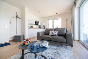 Un apartamento moderno con un sofá gris, una pequeña mesa de comedor con frutas, y una cocina al fondo.