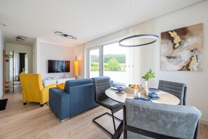 En moderne stue med sofa, stole, spisebord med morgenmad og et fjernsyn, samt et stort vindue med udsigt.