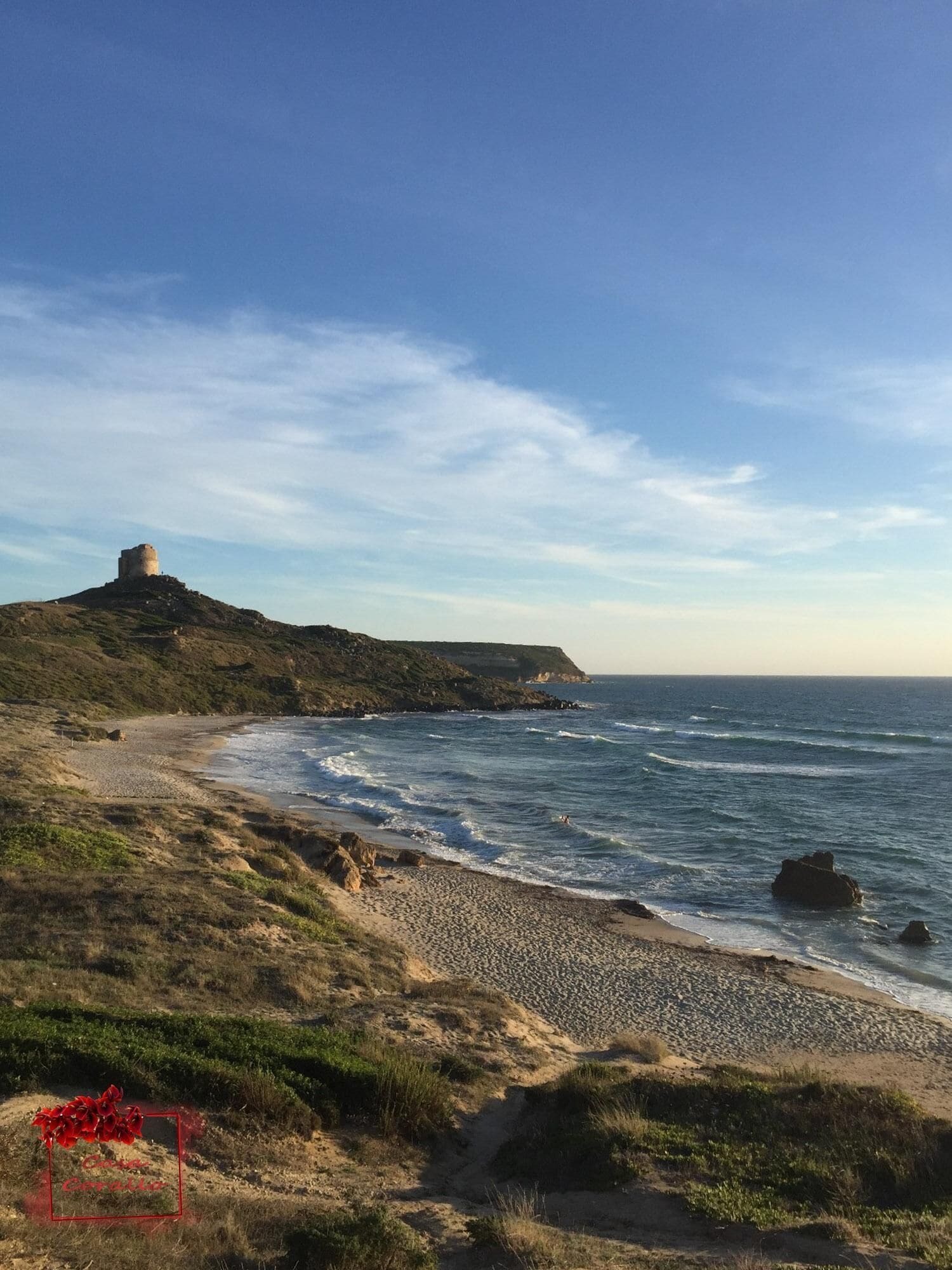 A coastal landscape with a hill featuring a historical structure, a pebble beach, and a clear sky.