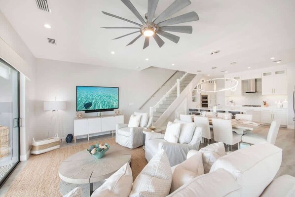 Ein modernes Wohnzimmer mit einem großen Flachbildfernseher, weißen Sofas, einem Essbereich und einer Treppe, die in den oberen Bereich führt.