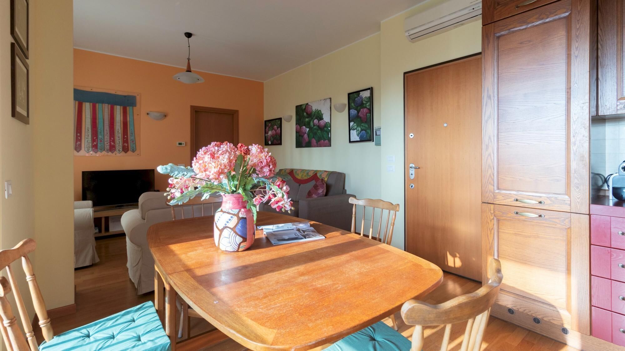 A dining area with a wooden table, chairs, and a vase of flowers. There is a television in the background and a door to the right.