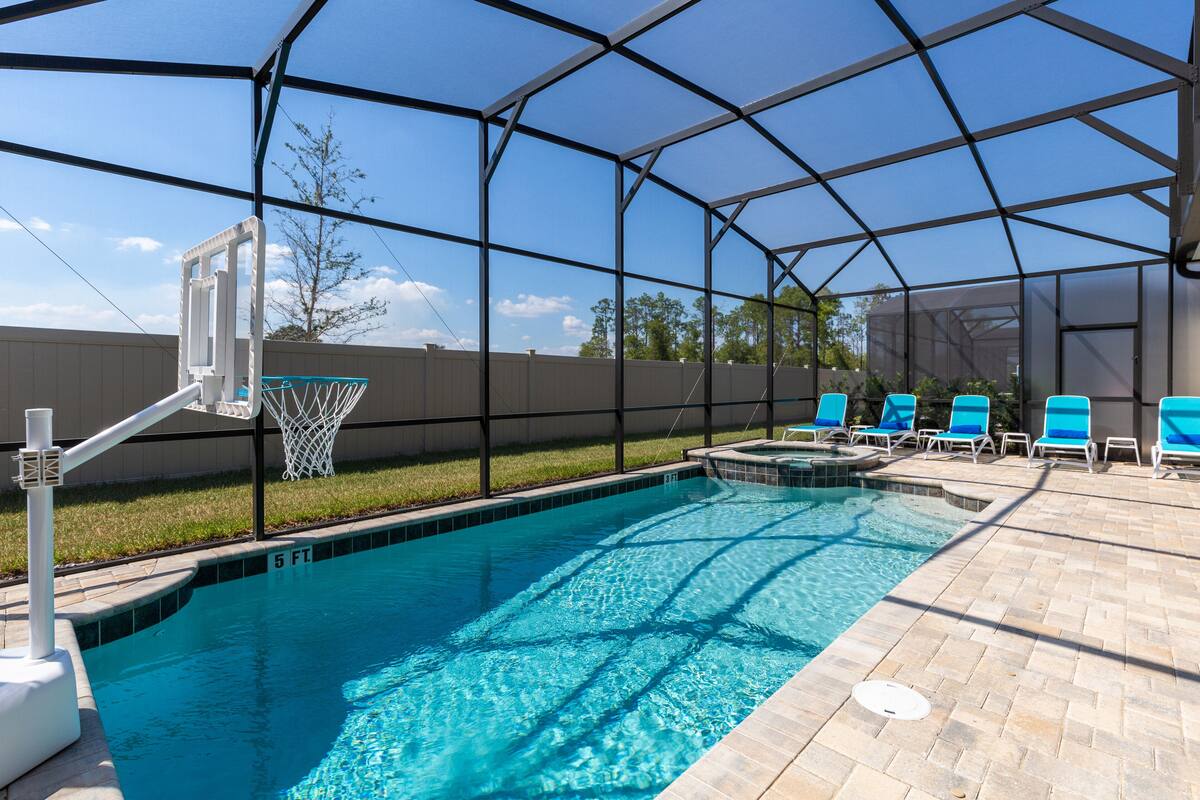 A swimming pool with a basketball hoop, lounge chairs, and a covered area.