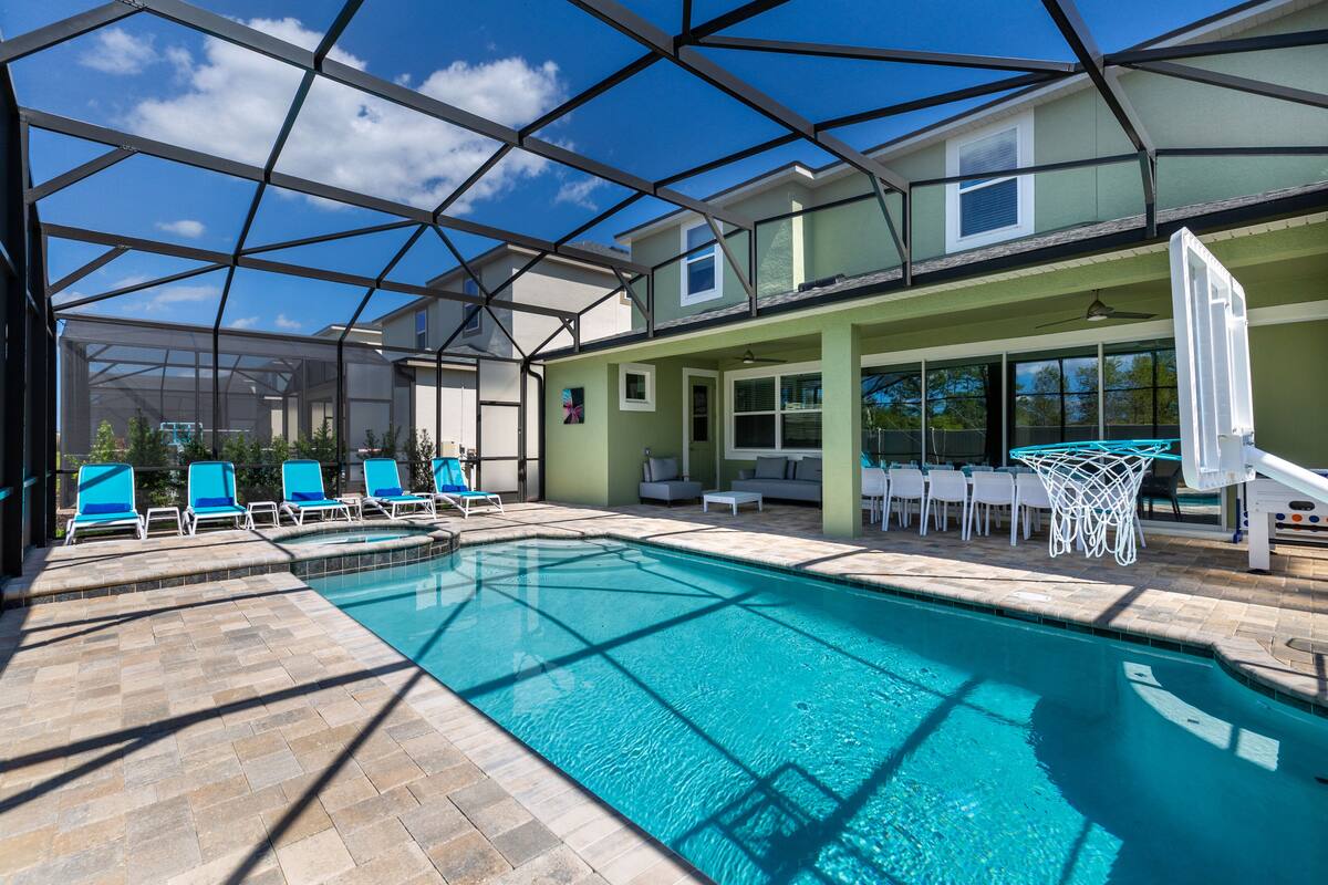 A swimming pool with lounge chairs and a basketball hoop, surrounded by a covered area with a view of the sky.
