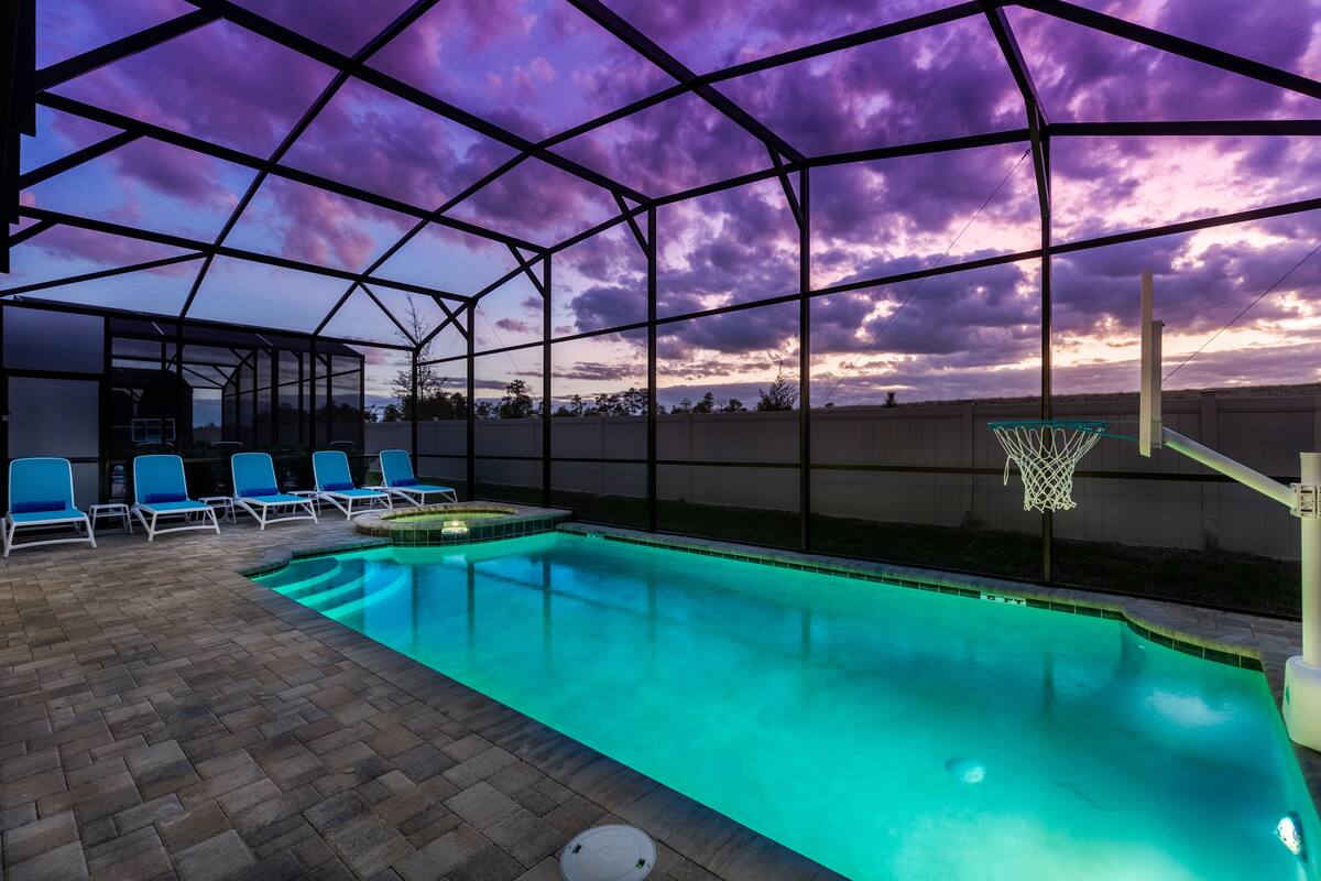 A swimming pool with a basketball hoop, surrounded by lounge chairs and a covered area with a metal frame.