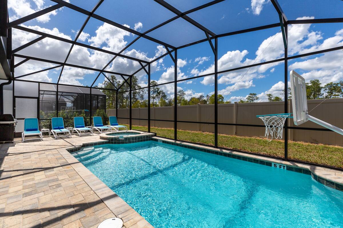 A swimming pool with a clear blue water, surrounded by a covered area with lounge chairs and a basketball hoop.