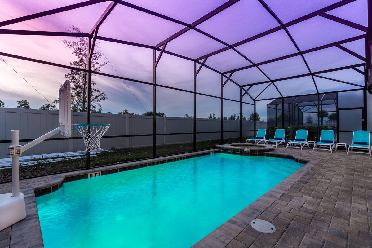 A swimming pool with a covered area, a basketball hoop, and lounge chairs.