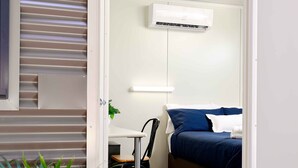 Habitación pequeña con cama, escritorio y silla. Hay una planta en maceta en la esquina.
