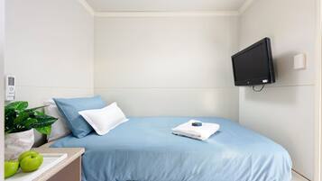 A small, neatly arranged bedroom with a bed, a TV mounted on the wall, a bedside table with a plant, and a window.