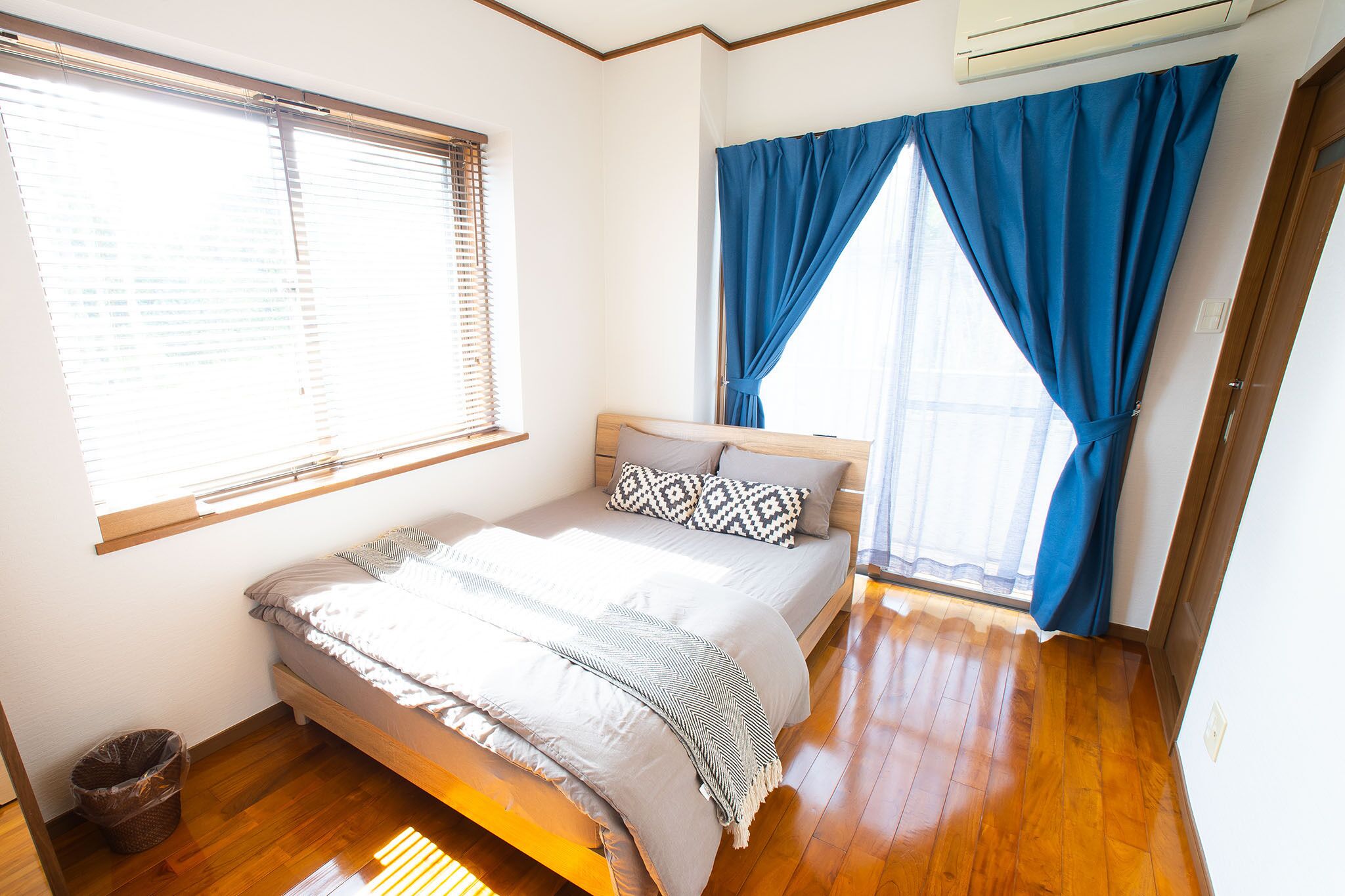 A bedroom with a bed, a window with blinds, a blue curtain, and a wooden floor.