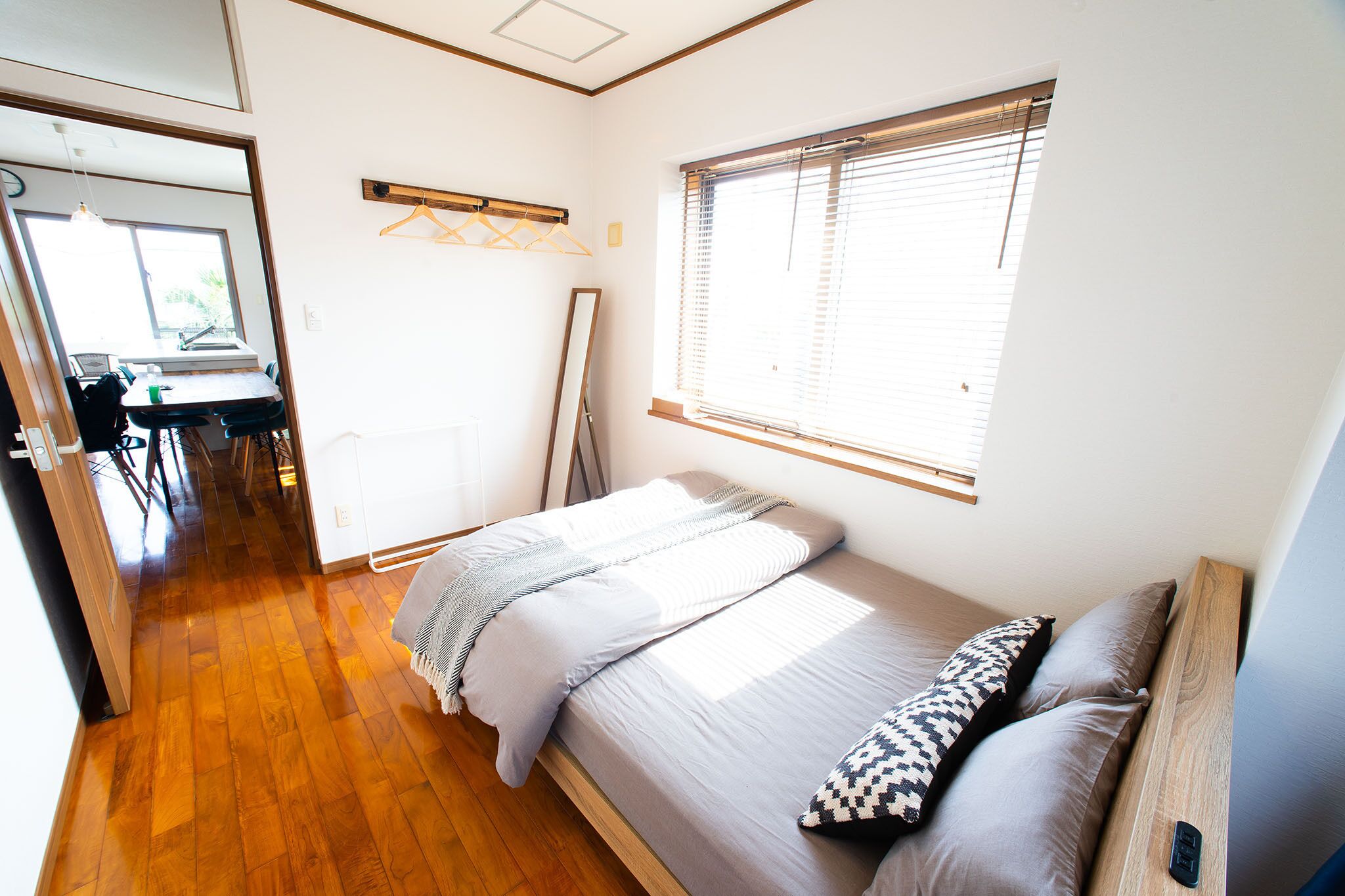 Un dormitorio con cama, ventana con persianas, suelo de madera y un pasillo que lleva a un baño.
