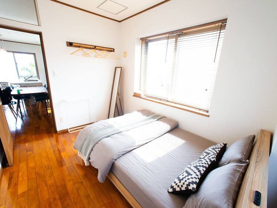 Un dormitorio con cama, ventana con persianas, suelo de madera y un pasillo que lleva a un baño.