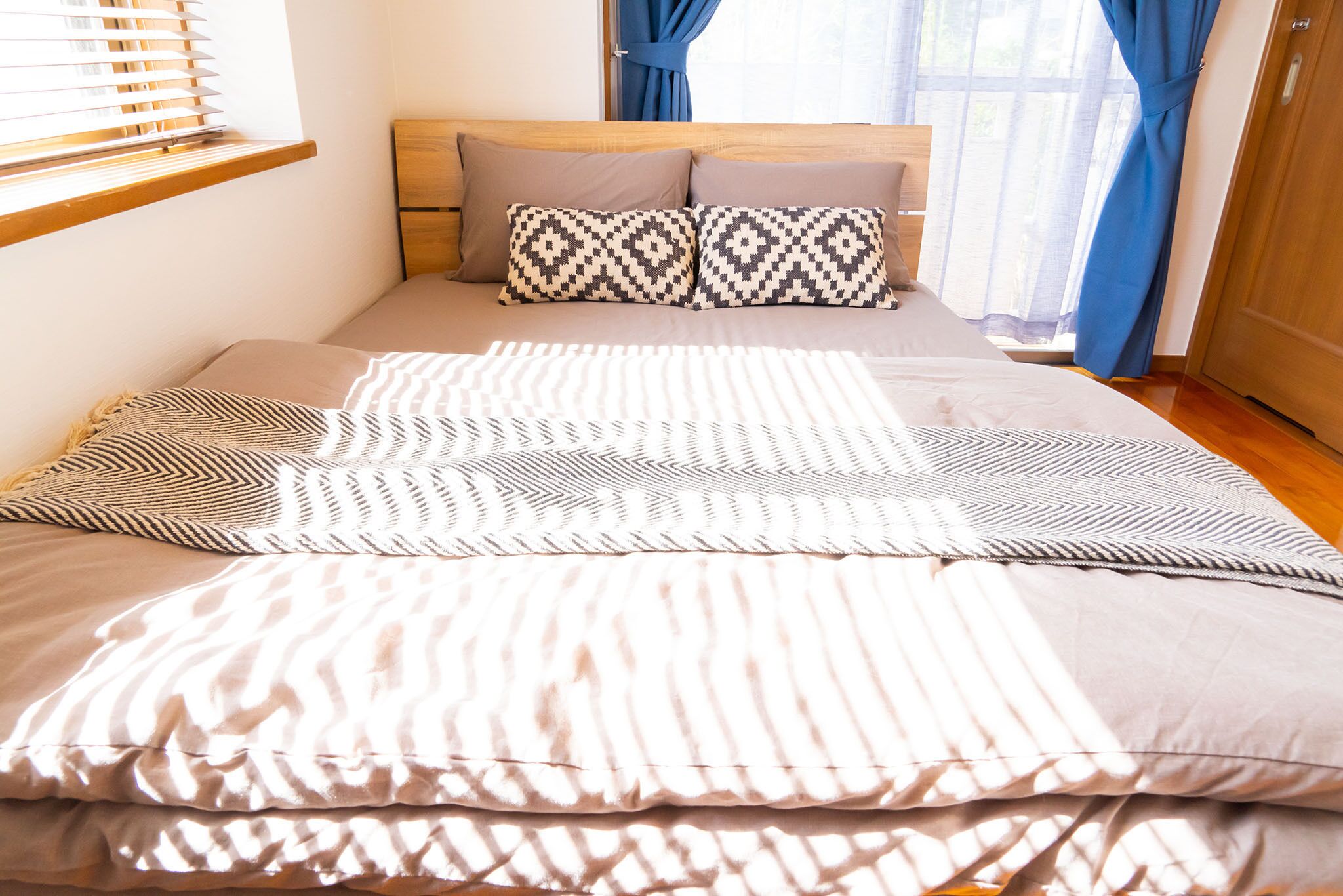 A neatly made bed with patterned pillows and a striped blanket, a wooden headboard, and a window with blinds.