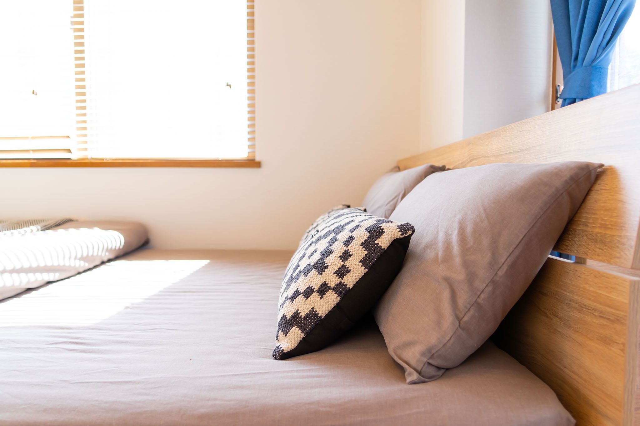 A neatly made bed with a patterned pillow and a plain pillow, a wooden headboard, and a window with blinds.