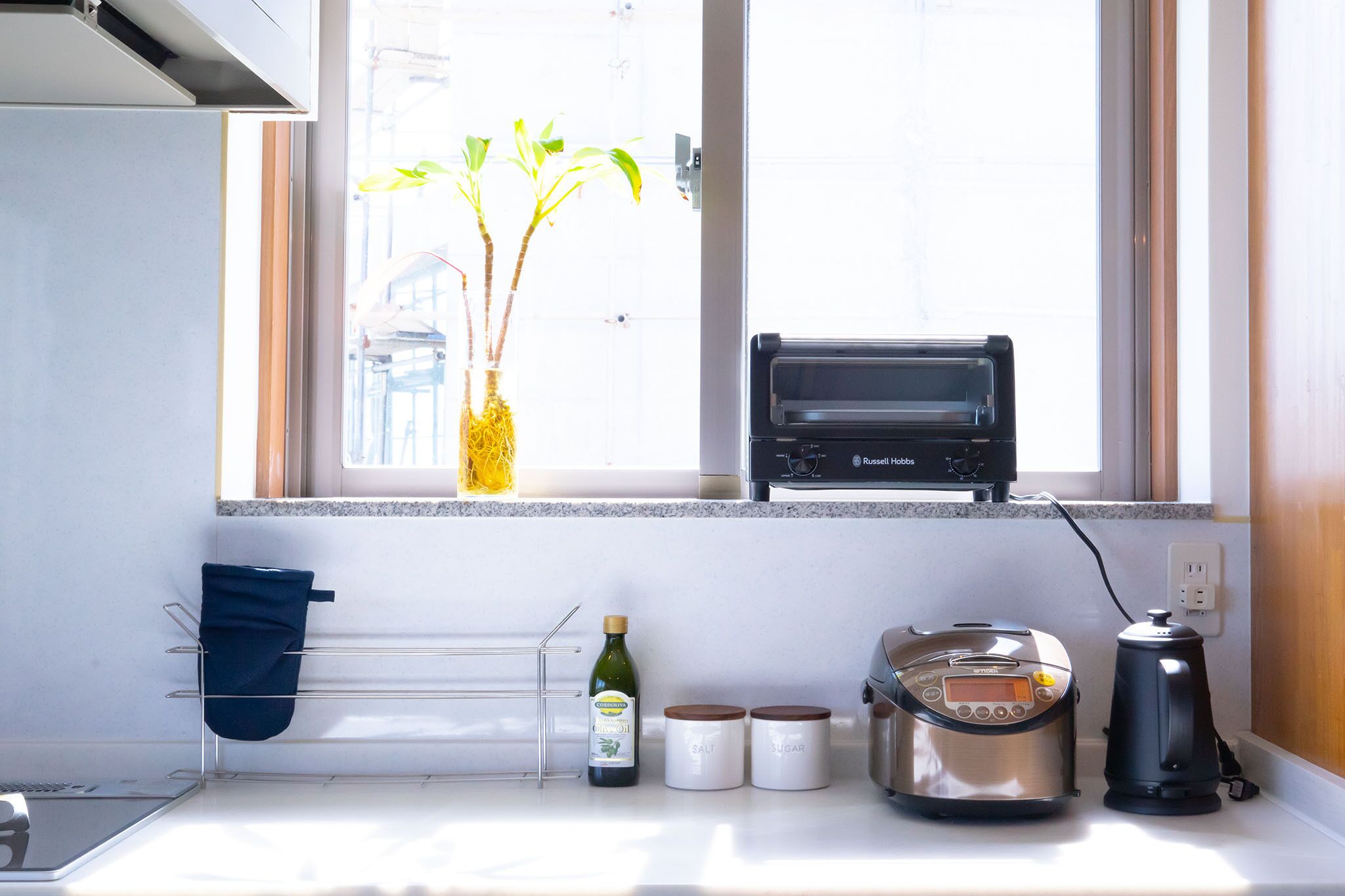 Una cocina con horno tostador, cocedor de arroz, recipientes de sal y azúcar, y una botella de aceite de oliva sobre la encimera.