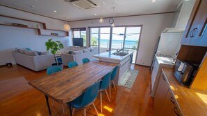 A modern kitchen with wooden flooring, a dining area with blue chairs, and a view of the ocean through large windows. - BLUE POINT OKINAWA (Yomitan)