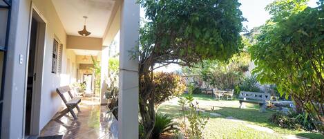A covered outdoor area with a bench, a pathway leading to a building entrance, and a garden with various plants and a tree.
