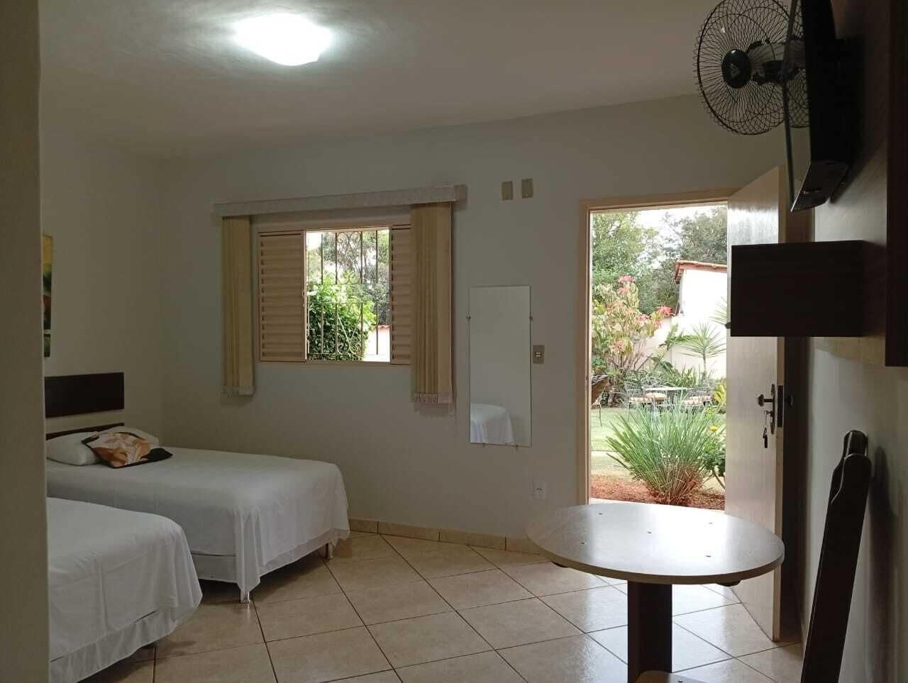 A room with two beds, a small table, a fan, and a window with blinds.