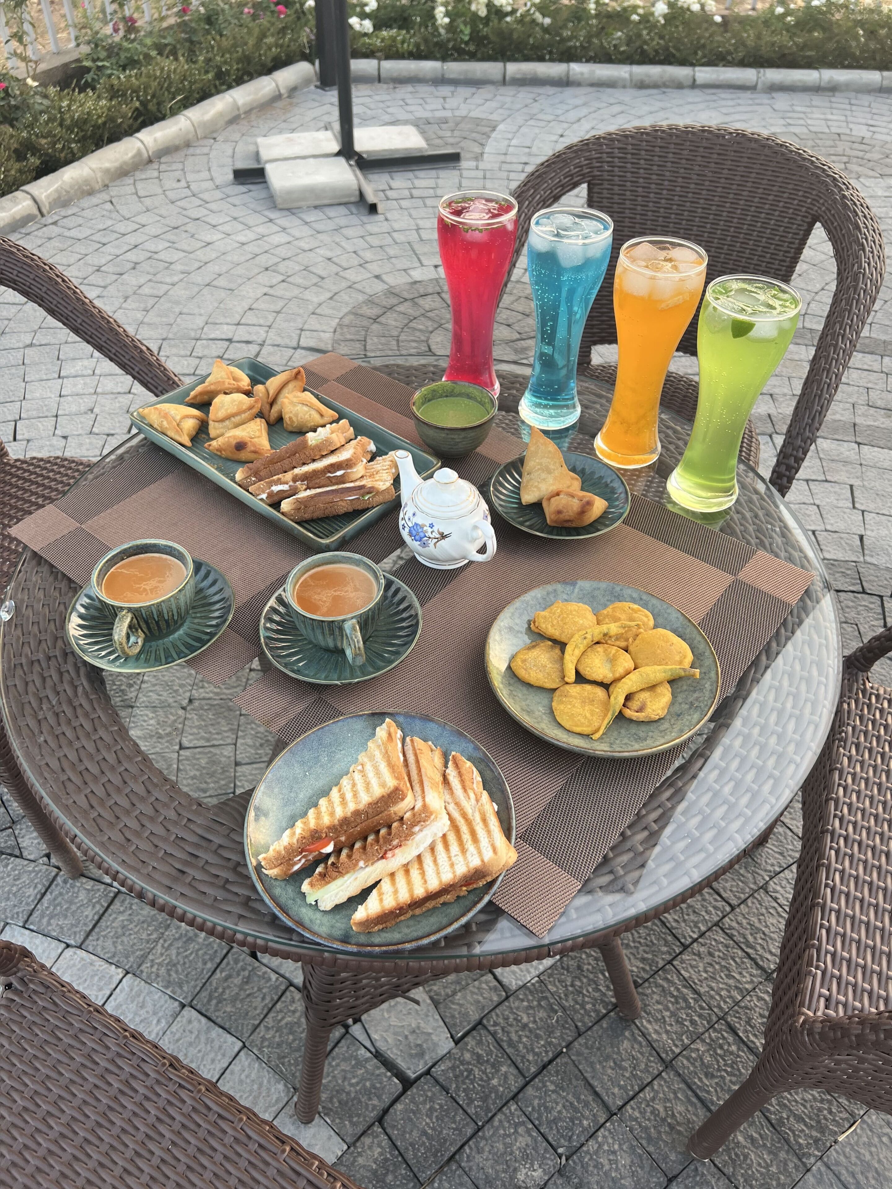 Une table ronde placée à l’extérieur, entourée de chaises en osier, avec des assiettes remplies de sandwichs, de pâtisseries et de boissons colorées.