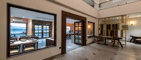 A spacious hotel lobby with large windows offering a view of the cityscape, a modern staircase, and a dining area with tables and chairs.