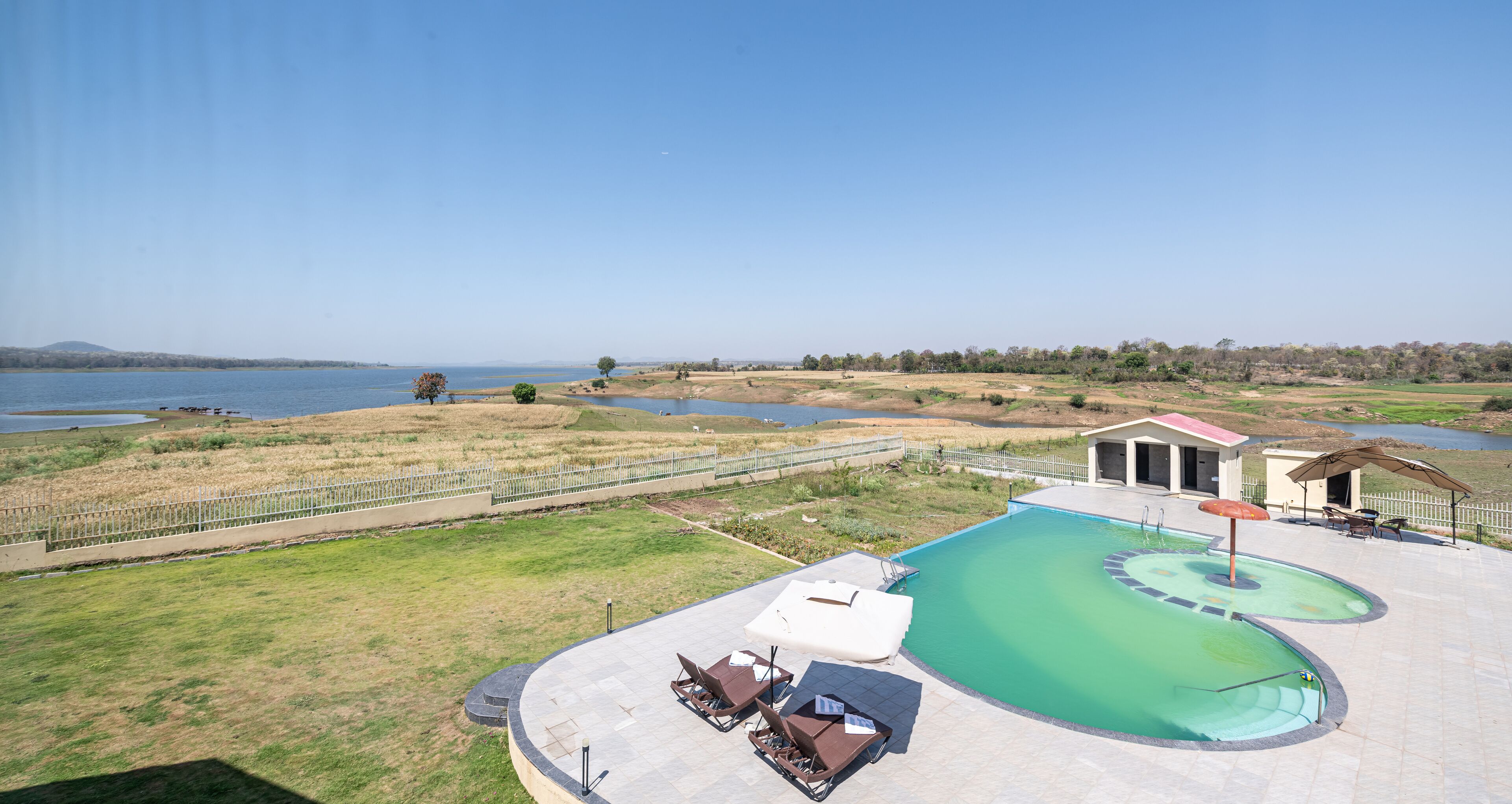 A swimming pool with lounge chairs and a small building, surrounded by a fenced area and a view of a lake.