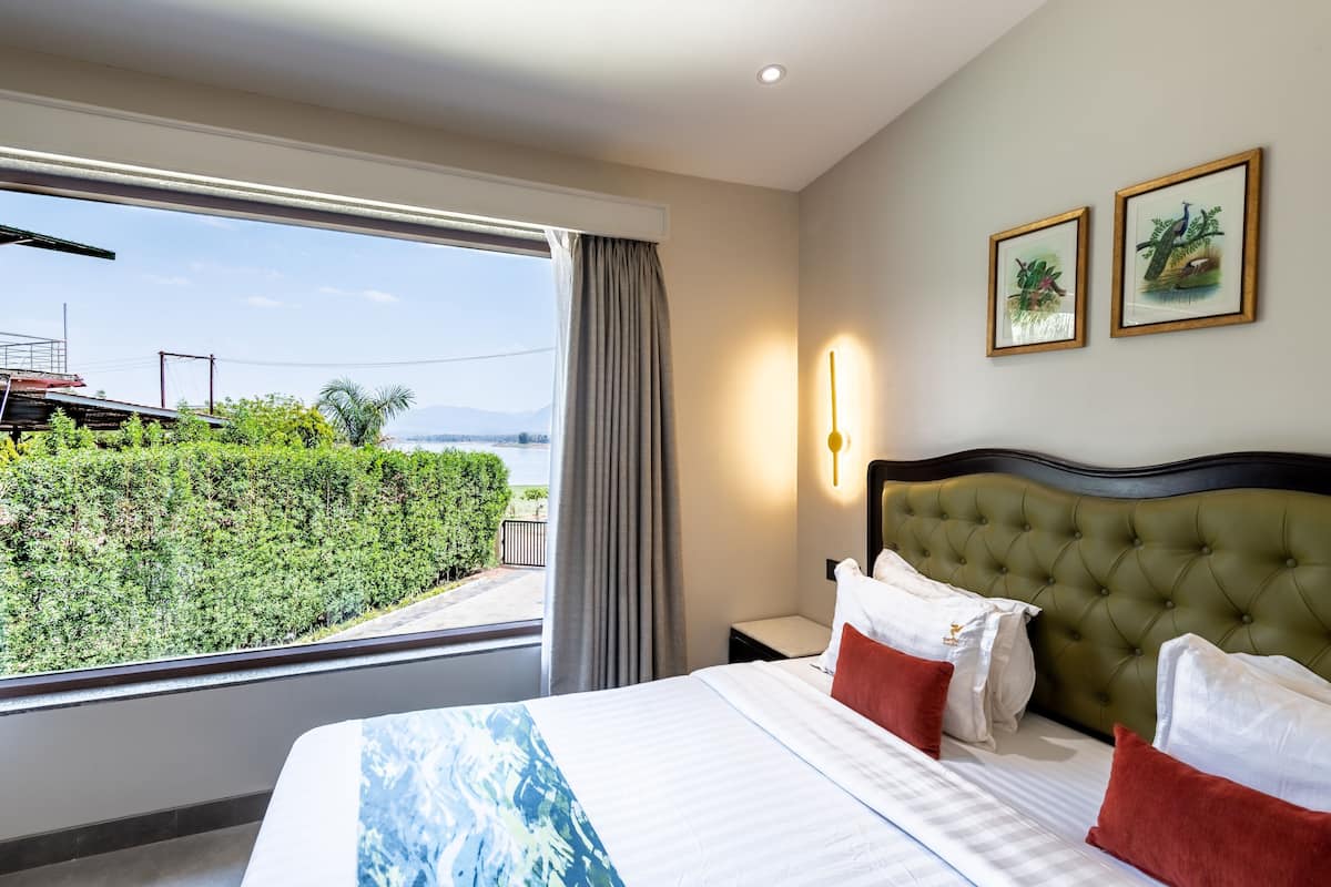 Quarto de hotel com uma cama grande, vista para vegetação e montanhas, e duas fotos emolduradas na parede.