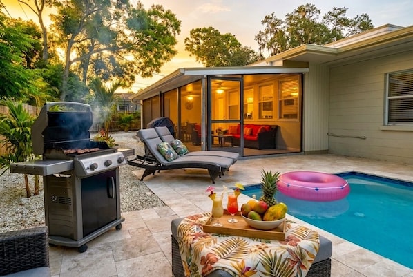 A backyard with a grill, lounge chairs, a pool, and a dining table with drinks and fruit.