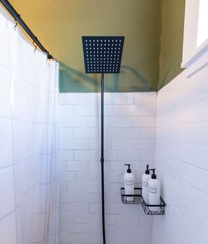 Une douche dotée d’un pommeau de douche à pluie noir, de murs recouverts de carreaux blancs et de deux bouteilles de shampoing posées sur une étagère noire.