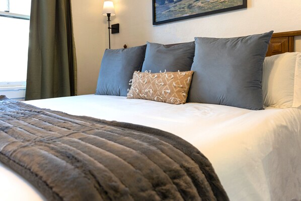 Un lit bien fait, avec une literie aux tons de gris et de blanc, un plaid à motifs et une lampe fixée au mur.