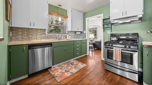 Une cuisine avec des meubles de couleur verte, un lave-vaisselle en acier inoxydable, une cuisinière et une fenêtre avec des stores.