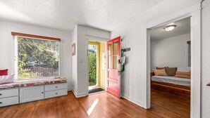 Une pièce avec un plancher en bois, un banc, une porte rouge et une fenêtre avec des stores.