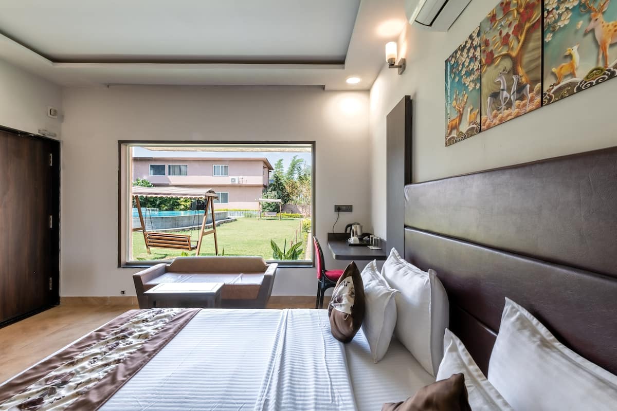 Quarto de hotel com uma cama grande, uma escrivaninha, uma cadeira, uma janela com vista para edifícios e vegetação, e uma obra de arte pendurada na parede.