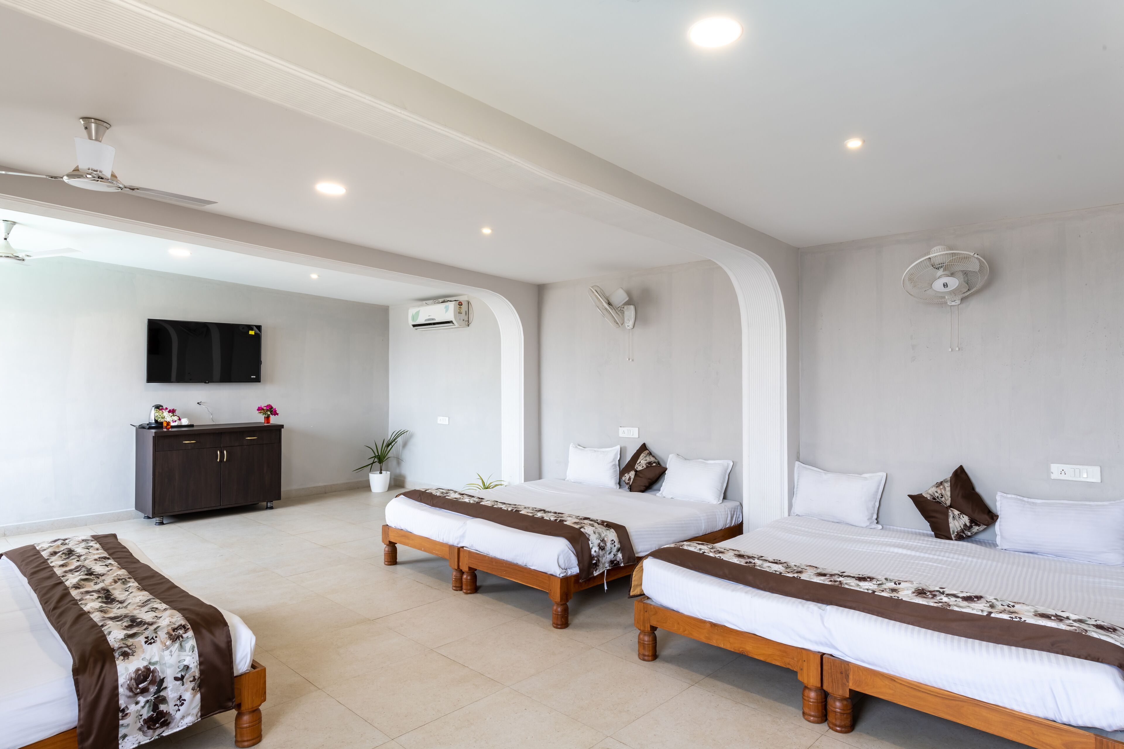 Habitación de hotel con tres camas, televisor, unidades de aire acondicionado y ventilador de techo.