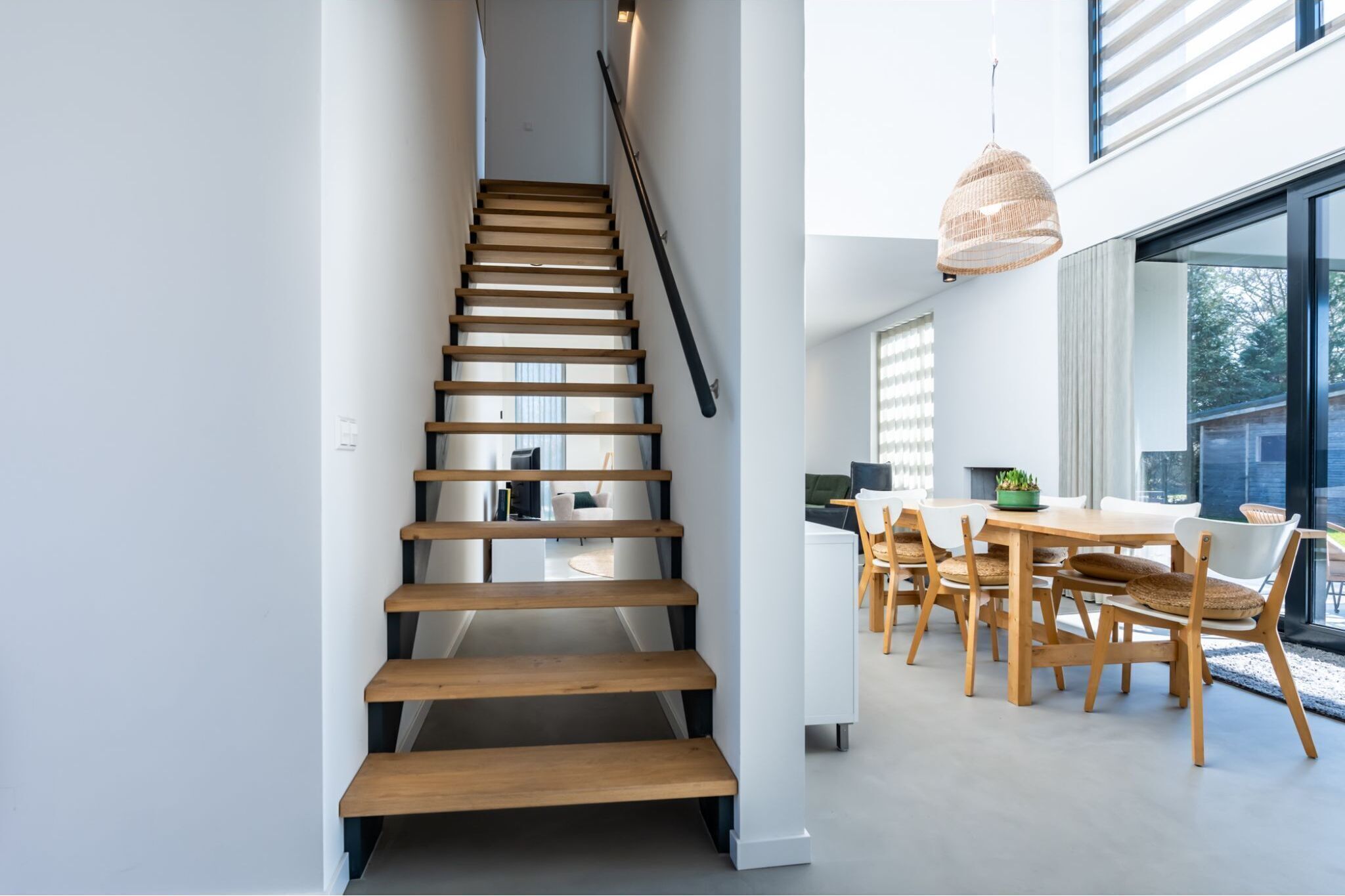 Een moderne trap met houten treden en een zwarte leuning, die leidt naar een eetgedeelte met een tafel en stoelen.