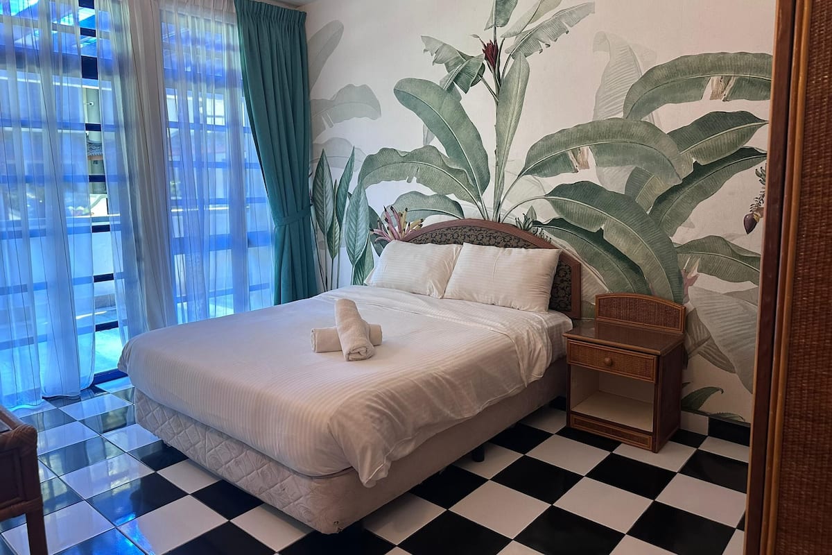 A neatly made bed with a white comforter and pillows, a small wooden nightstand with a lamp, and a large window with sheer curtains.
