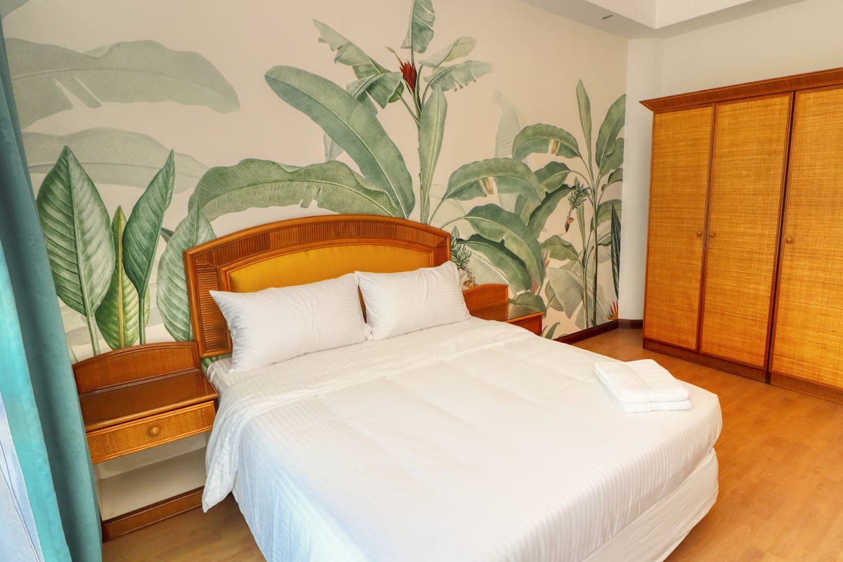 A bedroom with a bed, wooden wardrobe, and a wall with a tropical plant mural.