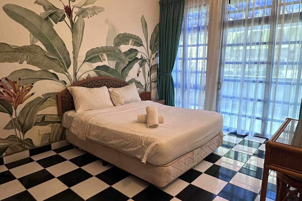 A hotel room with a checkered floor, a bed with white linens, a large plant mural on the wall, and a window with curtains.