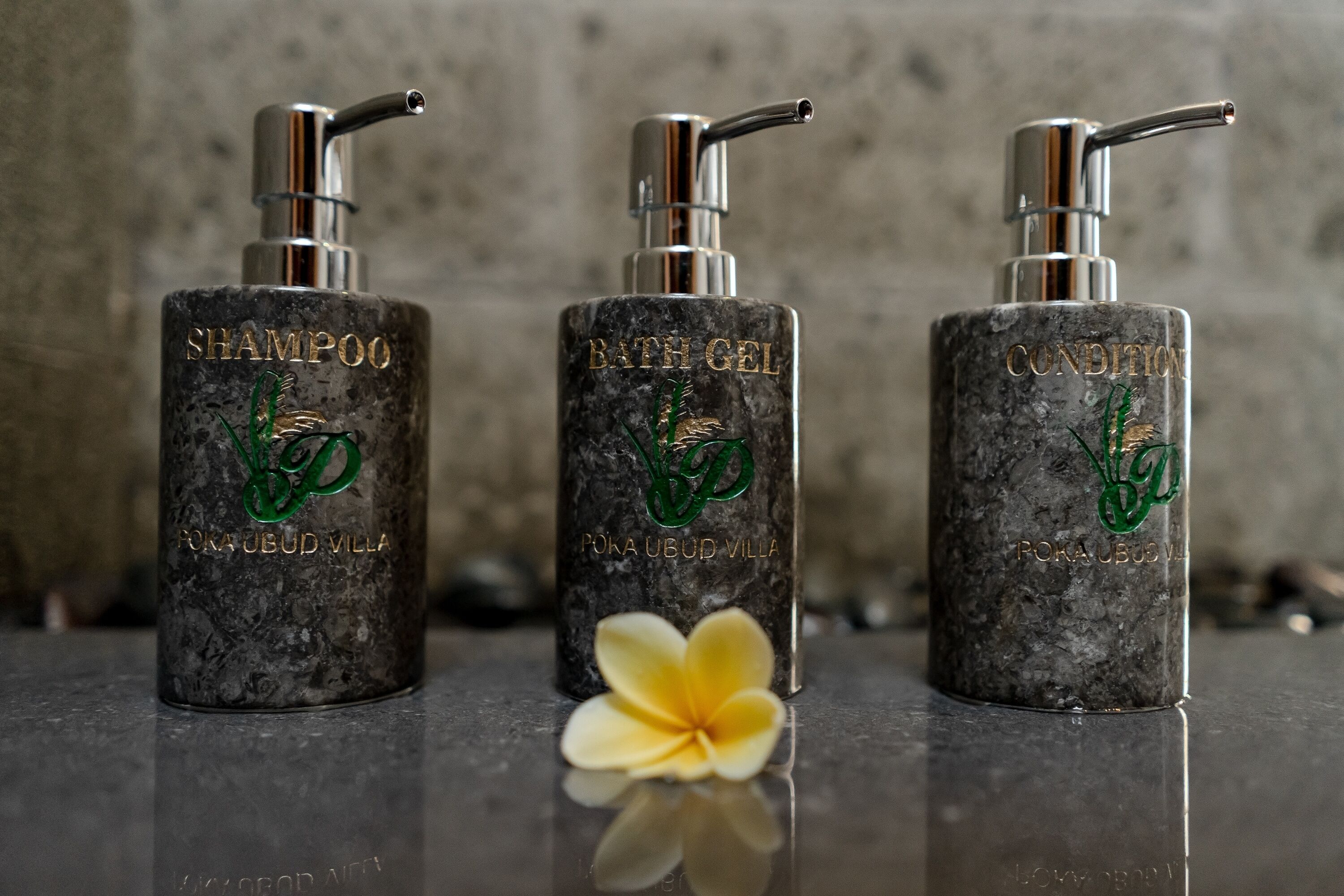 Three bottles of Poka Ubud Villa shampoo, bath gel, and conditioner on a reflective surface with a flower.