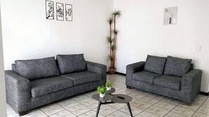 Una sala de estar con dos sofás grises, una mesita redonda con una planta y una planta alta en maceta contra la pared.