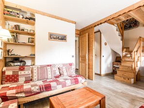A cozy living room with a wooden sofa, a coffee table, and a bookshelf filled with books and decorative items. A staircase leads to an upper level.