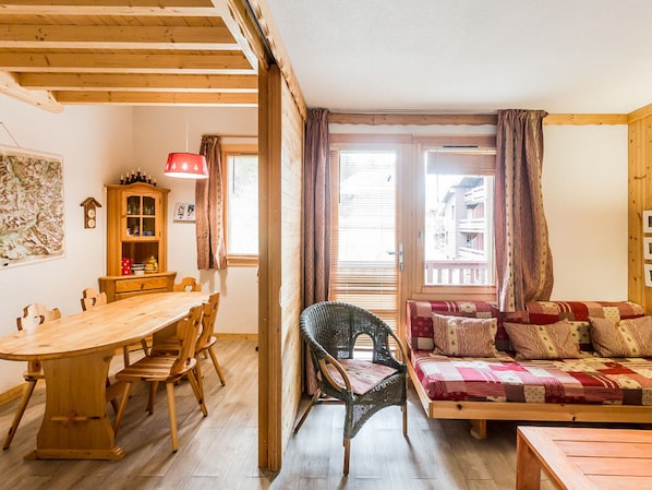 A cozy living room with a wooden dining table, chairs, a wicker chair, a sofa with patterned cushions, and a wooden cabinet.