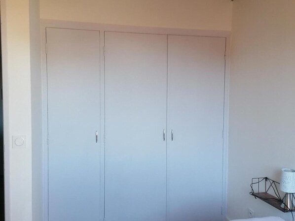 A bedroom with a large white wardrobe, a bedside table with a lamp, and a small shelf.