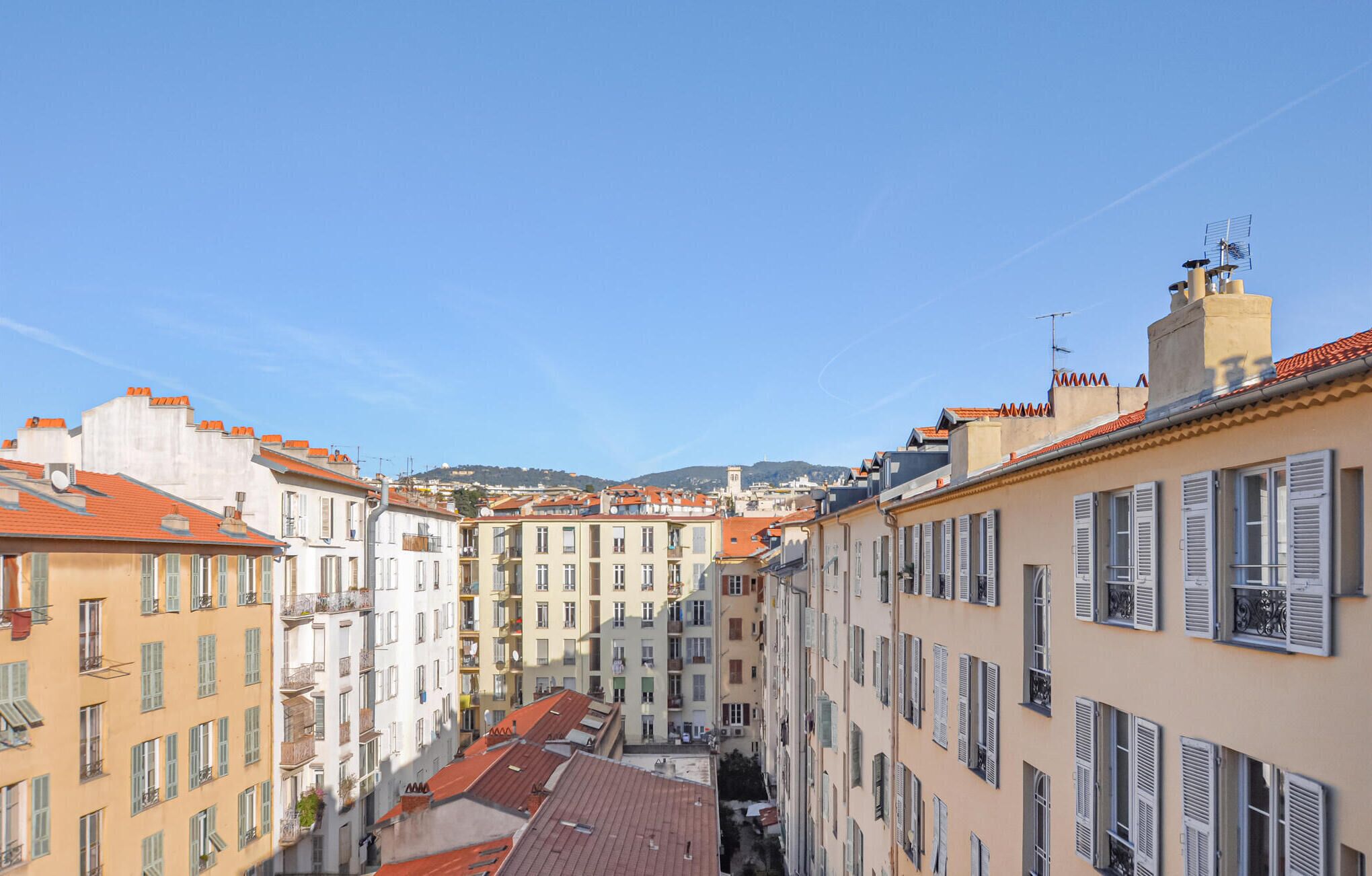 Una calle estrecha flanqueada por edificios de varios pisos, con balcones y tejados de tejas rojas.