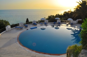A circular infinity pool with lounge chairs and umbrellas, overlooking a body of water at sunset. - Apartments Maria with Pool - Agios Gordios Beach, Corfu (Agios Gordios)