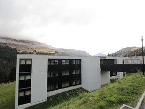 Ein modernes Gebäude mit flachem Dach und zahlreichen Fenstern, umgeben von einer grünen Fläche und Bergen im Hintergrund.