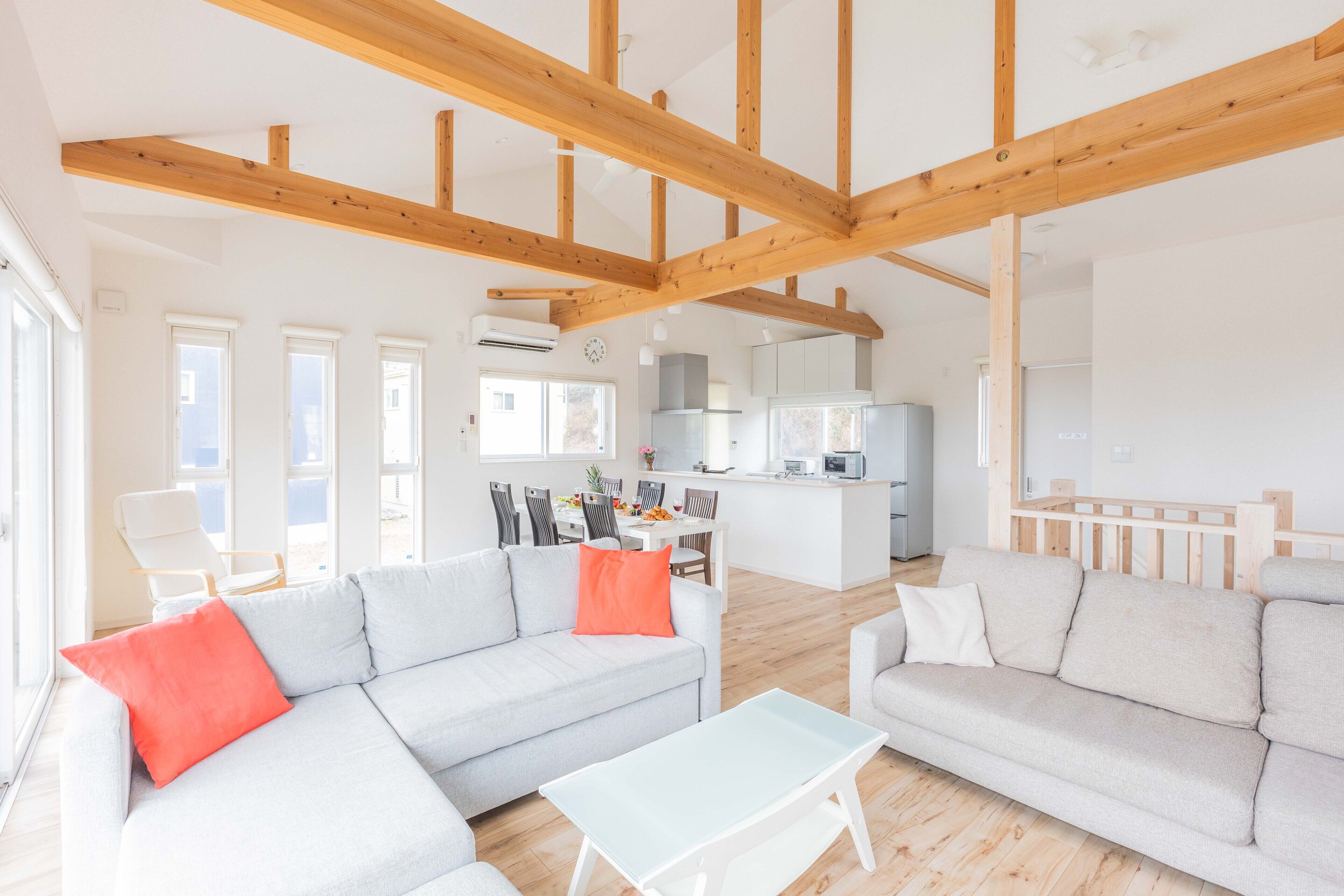 A spacious living area with a high ceiling and exposed wooden beams, a light grey sofa with red cushions, a white coffee table, and a kitchen in the background.