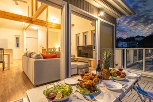 Una mesa de comedor dispuesta con comida y bebidas, un sofá y un televisor en un salón moderno con vigas de madera y un balcón con vistas a la ciudad.