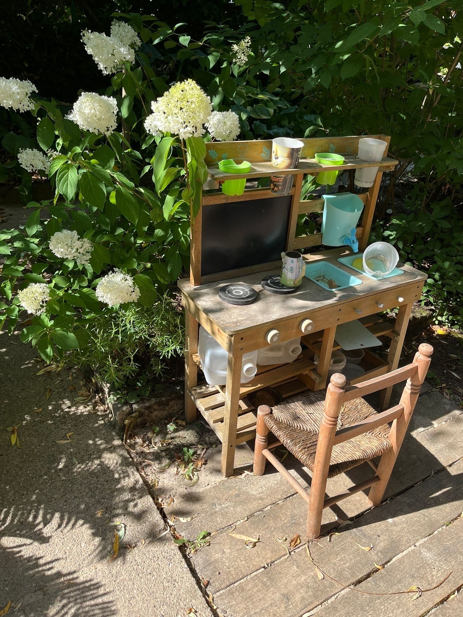 Una mesa de madera rústica al aire libre con una silla, rodeada de follaje y flores blancas.
